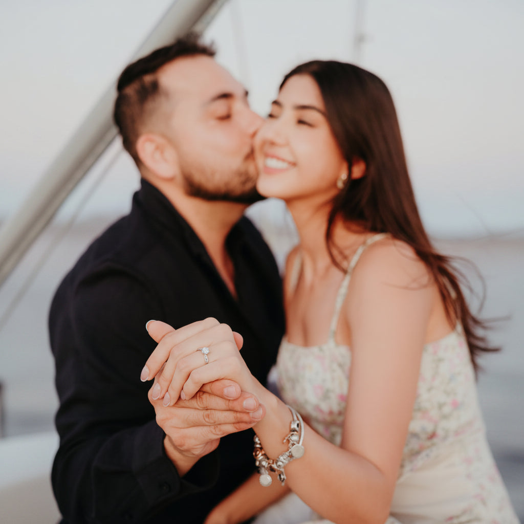 A couple embracing on a boat, with the man kissing the woman's cheek and her holding his hand. She is wearing a light floral print spaghetti-strap dress and silver bracelets, and he is wearing a black shirt.